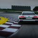 rear view of the 2023 audi r8 gt in grey as it makes a left-hand turn around a curve on a race track. the audi is crawling up on the red and white striped curb of the track, the sky is dusky in the background